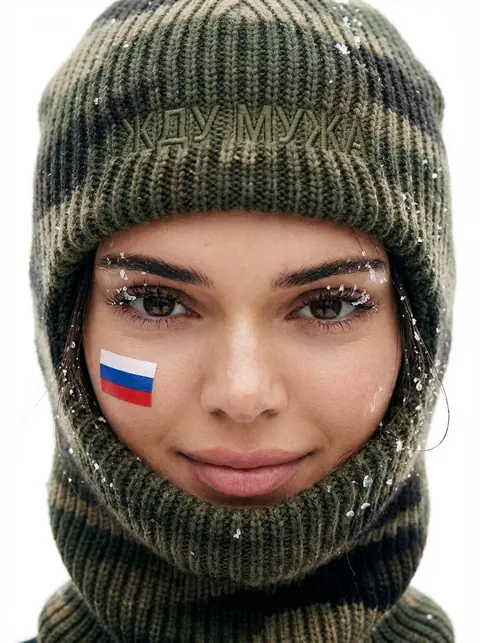 Промт модное фото через ИИ с женщиной в камуфляжной вязаной балаклаве на 23 февраля ЖДУ МУЖА и флагом России на щеке